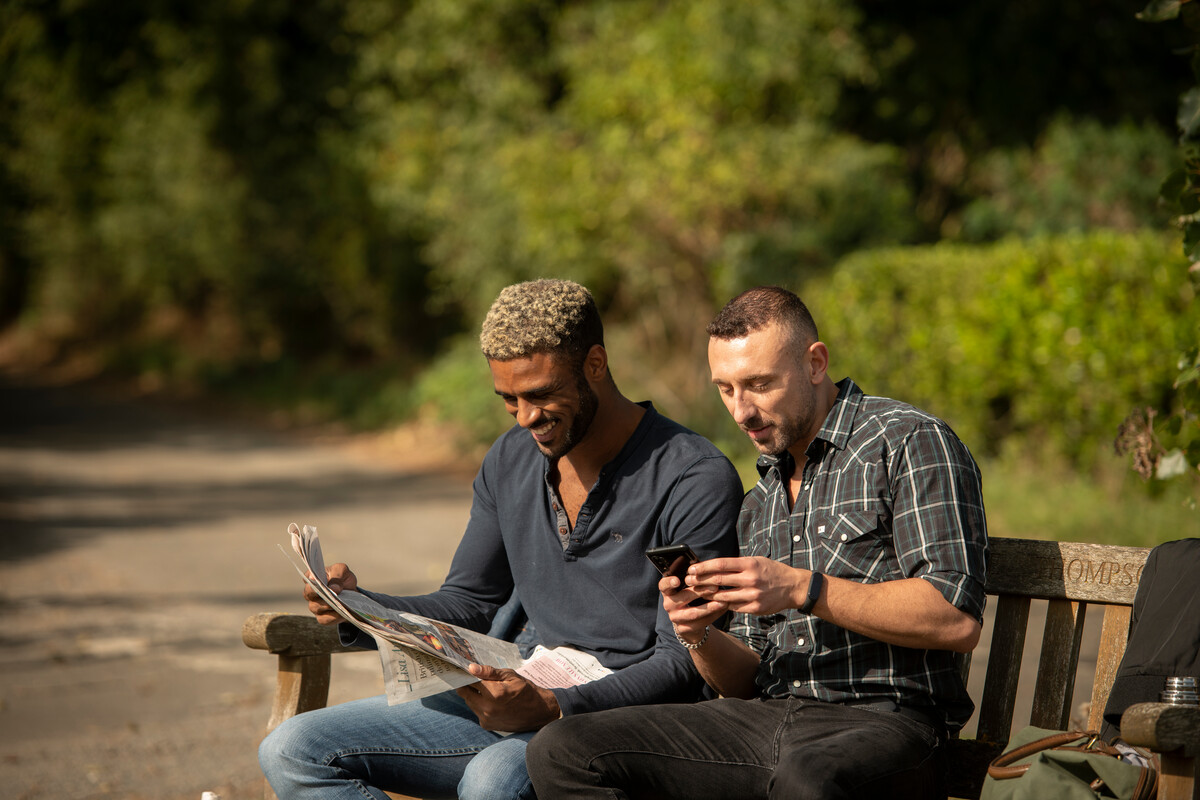 Two men sat on a bench in the countryside reading the news in a newspaper and on their phone.
