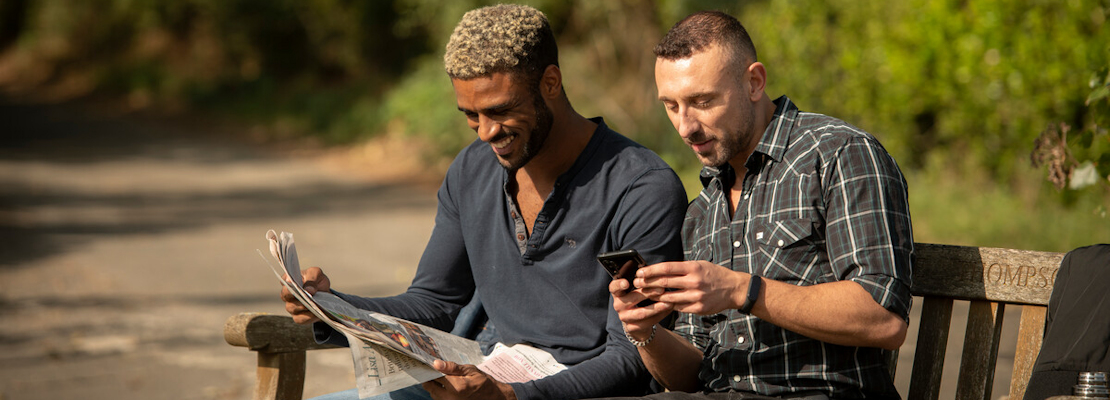 Two men sat on a bench in the countryside reading the news in a newspaper and on their phone.