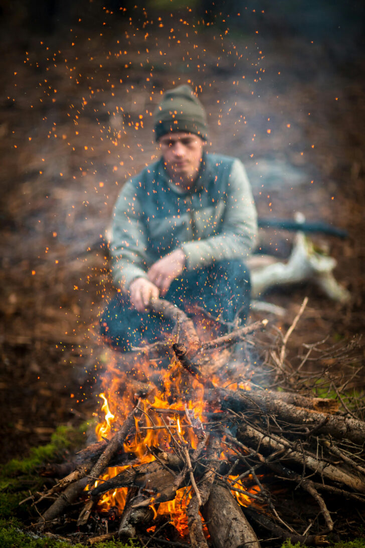 A man crouching behind a burning campfire