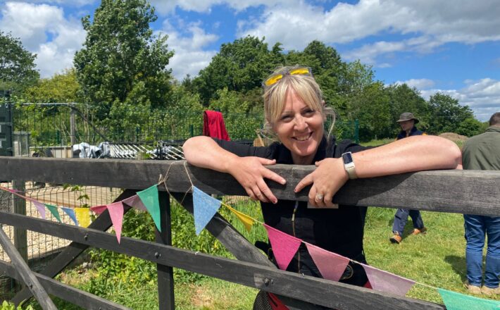 A woman smiling and leaning over a wooden 5-bar gate at Martha's Meadow