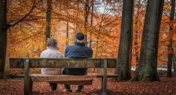 An older couple sitting on a bench enjoying the Autumn woodland in front of them.