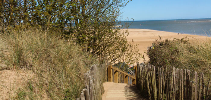 The view down the boardwalk path from the sand dunes to the expansive beach of golden sands below. The sea stretches off into the distance.