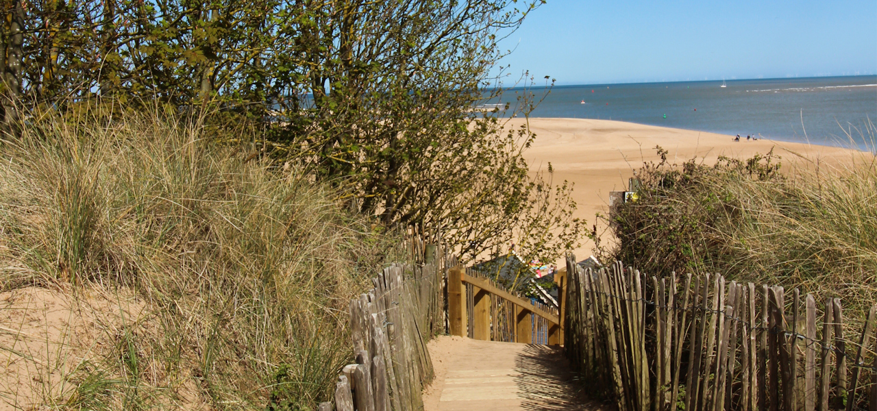 The view down the boardwalk path from the sand dunes to the expansive beach of golden sands below. The sea stretches off into the distance.