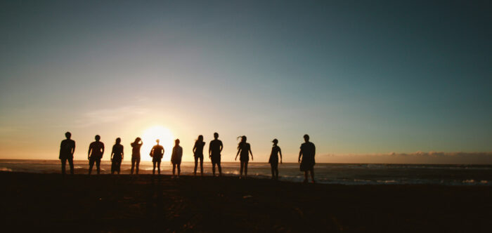 Silhouettes of a row of people standing facing away from the camera and watching the sun set over the sea on the horizon.