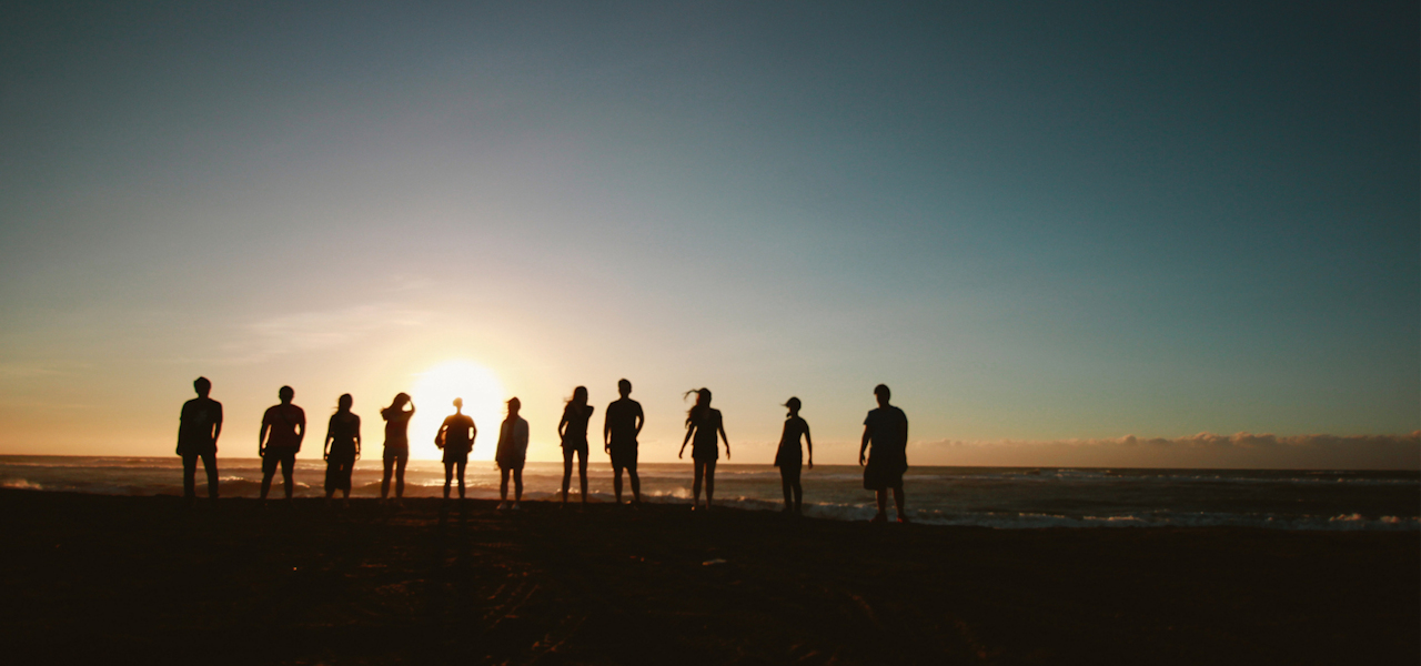 Silhouettes of a row of people standing facing away from the camera and watching the sun set over the sea on the horizon.
