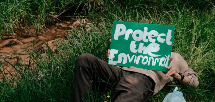 A man lies on his back in long grass next to a small stream. He is holding up a hand-painted sign saying 'Protect the Environment'.