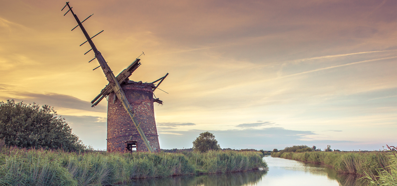 A derelict brick wind pump standing next to a reed-lined river which winds to the horizon.