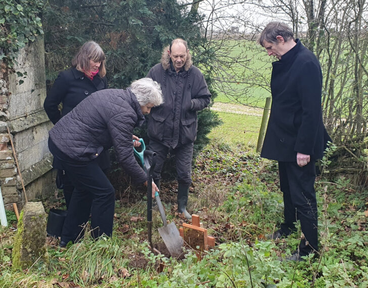Ian's wife holds a spade and plants a memorial silver birch tree, watched by three other people.